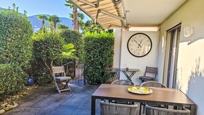 An outdoor patio with a table and chairs, surrounded by greenery and a clock on the wall. - Collina Verde B by Interhome (Ascona)