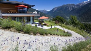 A modern house with a glass facade, outdoor seating, and a mountain view. - Apartment Zenkei by Interhome (Leontica)