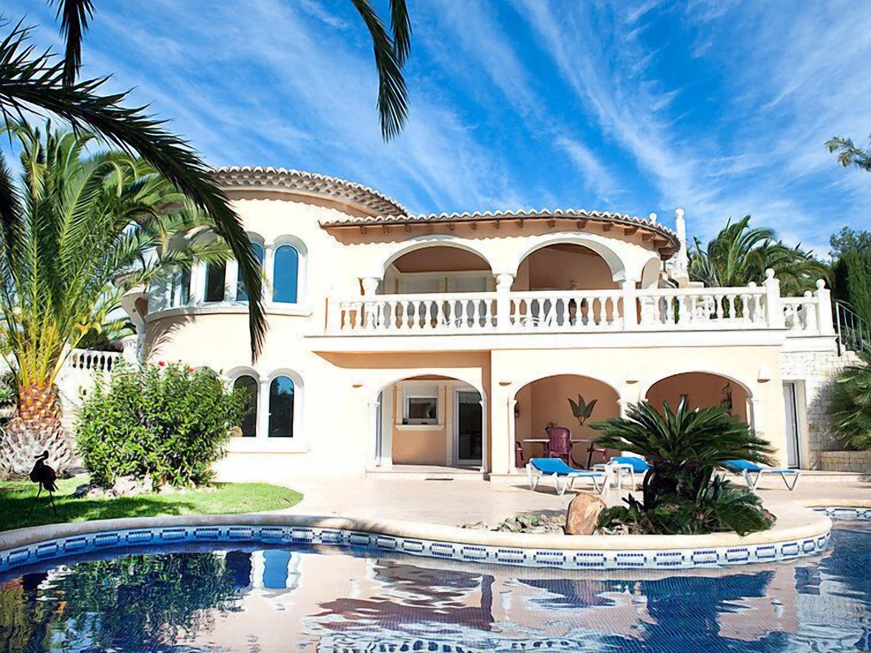 A large, two-story house with a swimming pool, surrounded by palm trees and a clear blue sky.