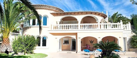 A large, two-story house with a swimming pool, surrounded by palm trees and a clear blue sky.