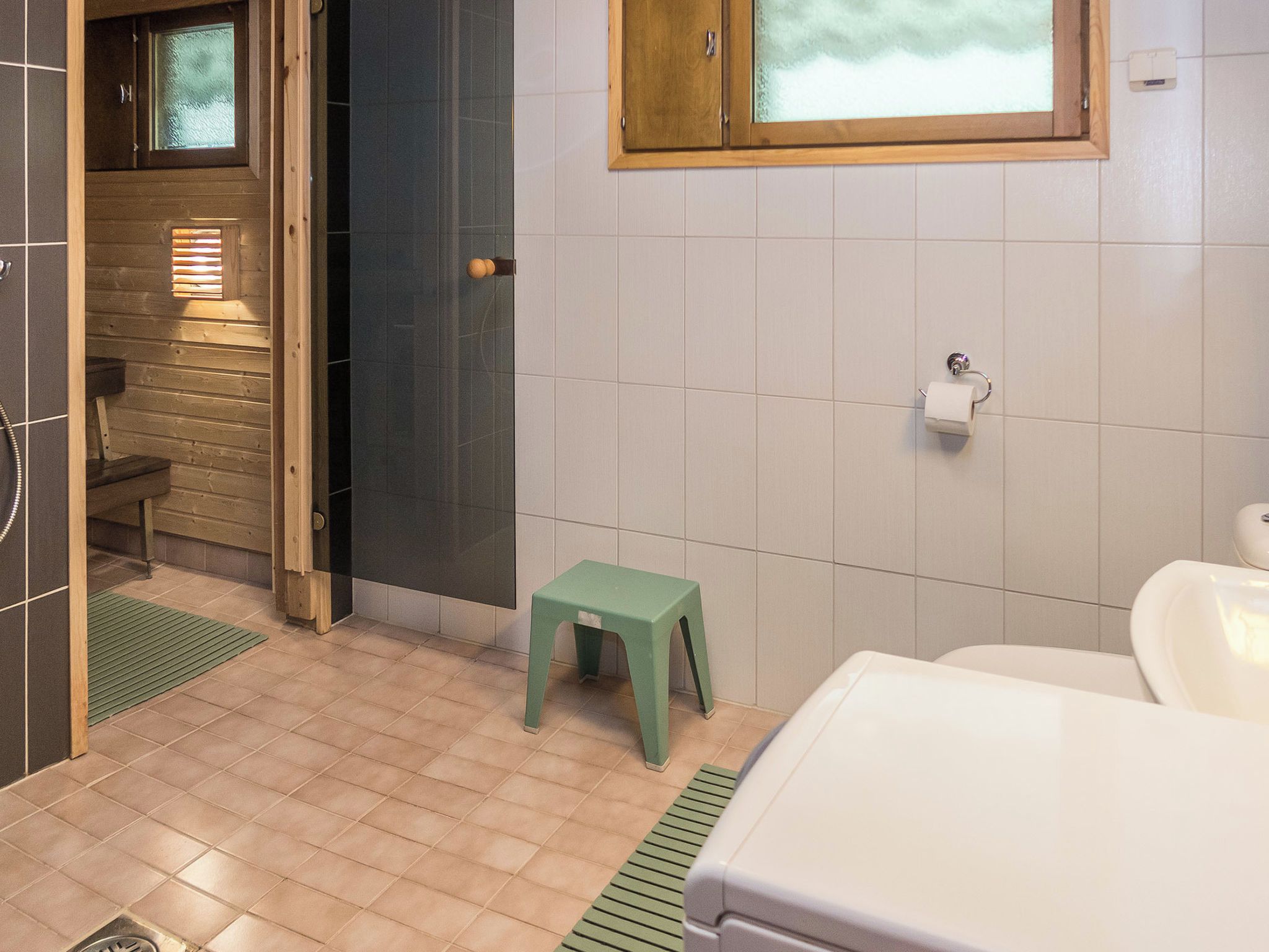 A bathroom with a toilet, a small stool, a window, and a towel dispenser.