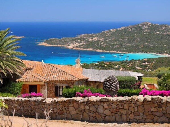 A stone-walled building with a tiled roof overlooking a coastal area with boats and a clear blue sea.