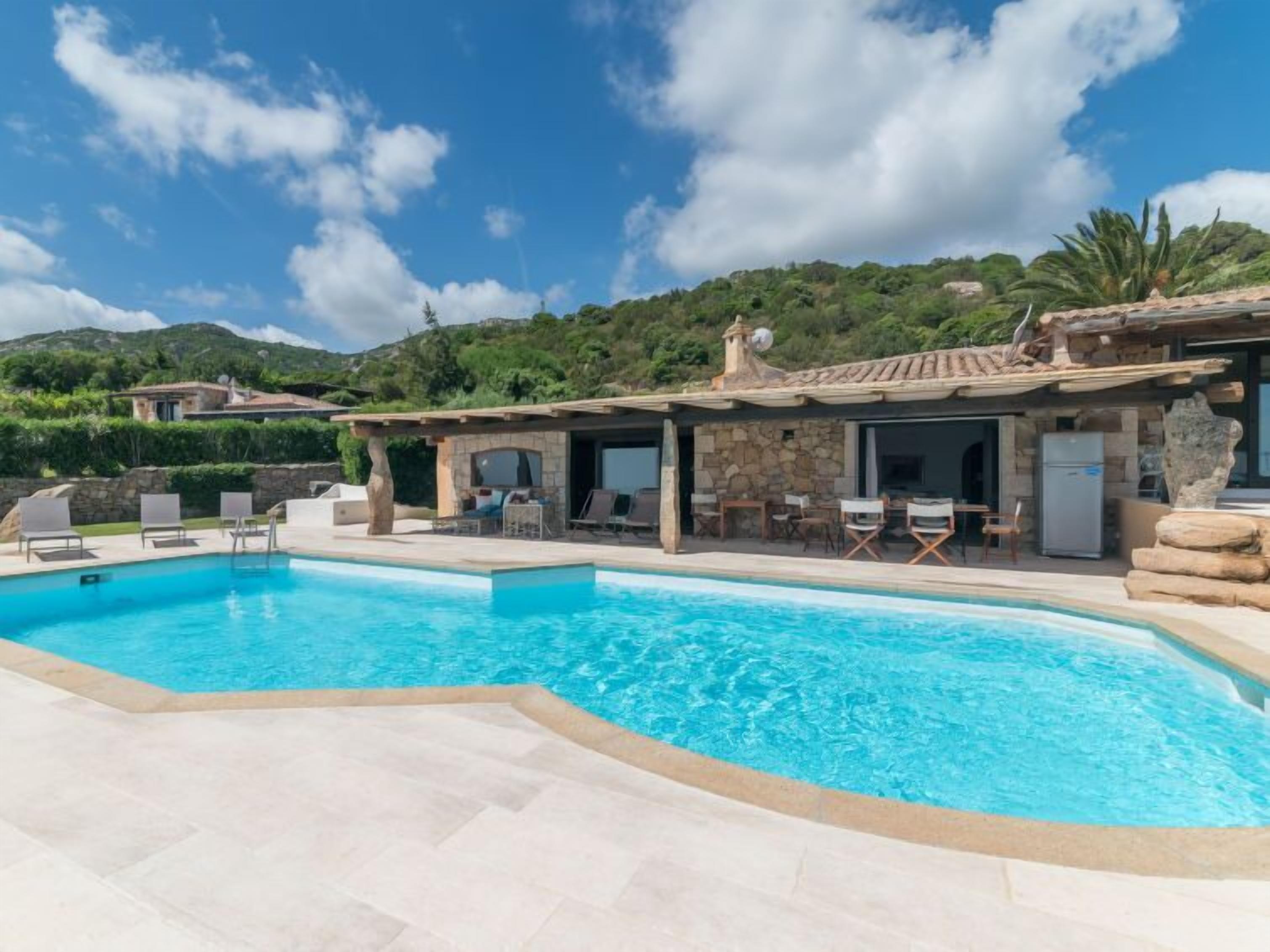 A poolside area with a clear blue pool, outdoor seating, and a stone building with a tiled roof.