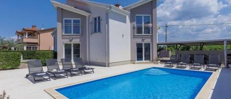 A modern two-story house with a swimming pool, sun loungers, and a covered patio area.