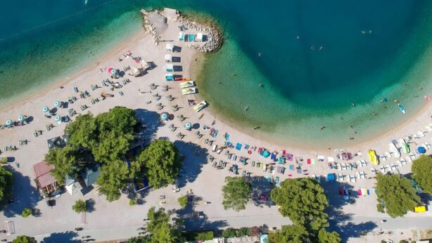 An aerial view of a beach with rows of sun loungers and umbrellas, a small rocky area, and clear blue waters.