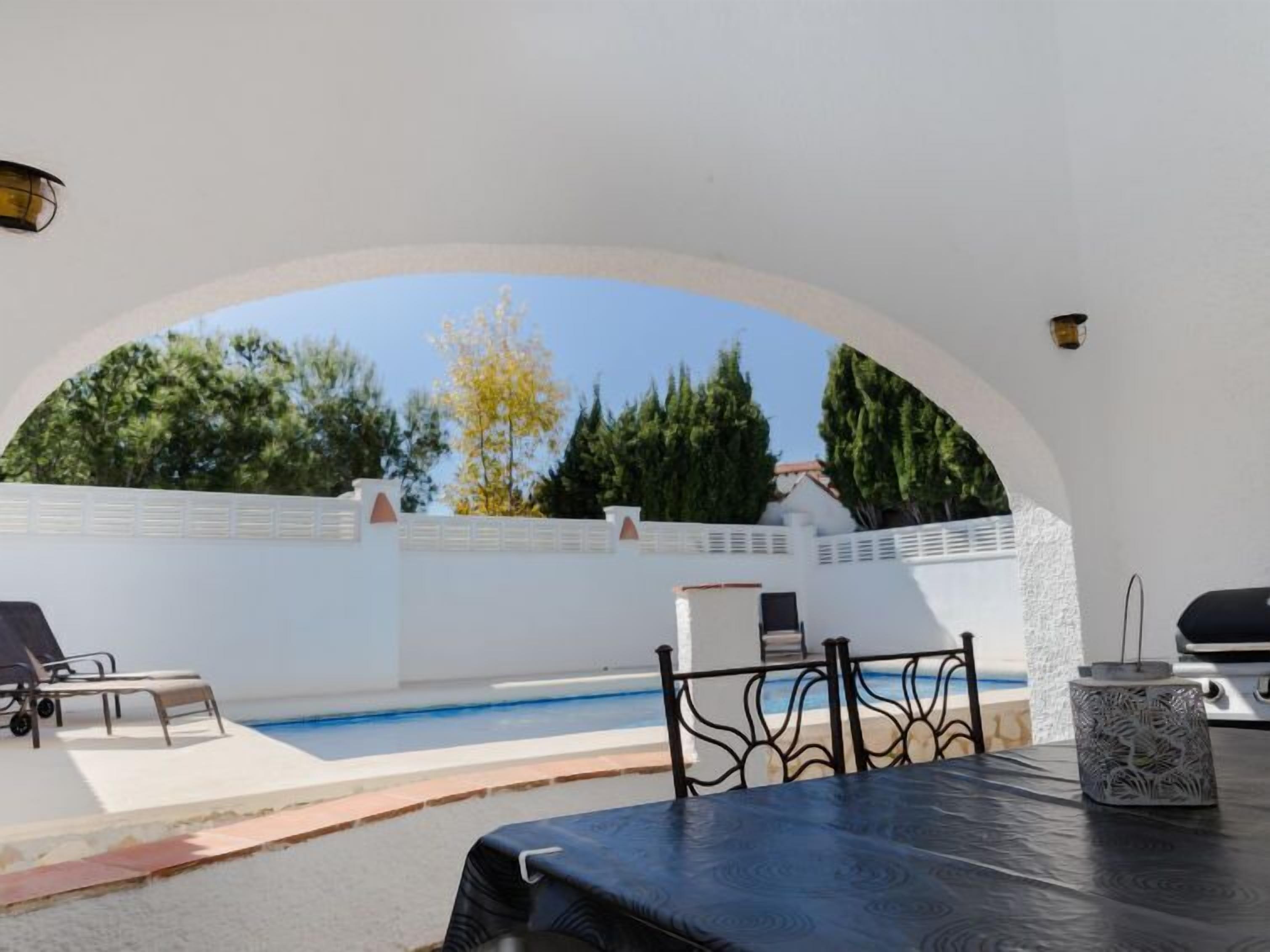Un espace extérieur avec une piscine, des chaises longues et une table, le tout entouré de murs blancs et d’arbres.