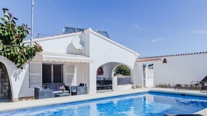 Zona de piscina con un edificio blanco, mobiliario exterior y un cielo azul claro.