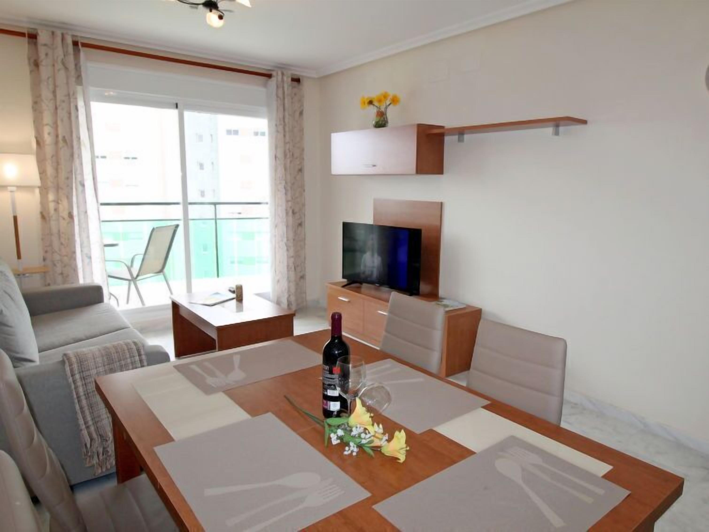 A modern living room with a wooden dining table, a sofa, a TV on a stand, and a balcony with a chair.