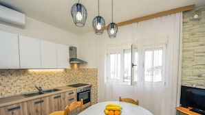 Une cuisine moderne avec des armoires blanches, un pare-feu en carreaux et un coin repas comprenant une table ronde et des chaises. On trouve un panier de fruits sur la table et un téléviseur posé sur un meuble.