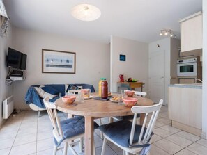 A cozy dining area with a round wooden table, white chairs, and a blue sofa. A television is mounted on the wall, and a framed picture hangs above the table.