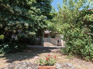 Un bâtiment en pierre avec un escalier vert menant à une porte, le tout entouré d’un feuillage dense.