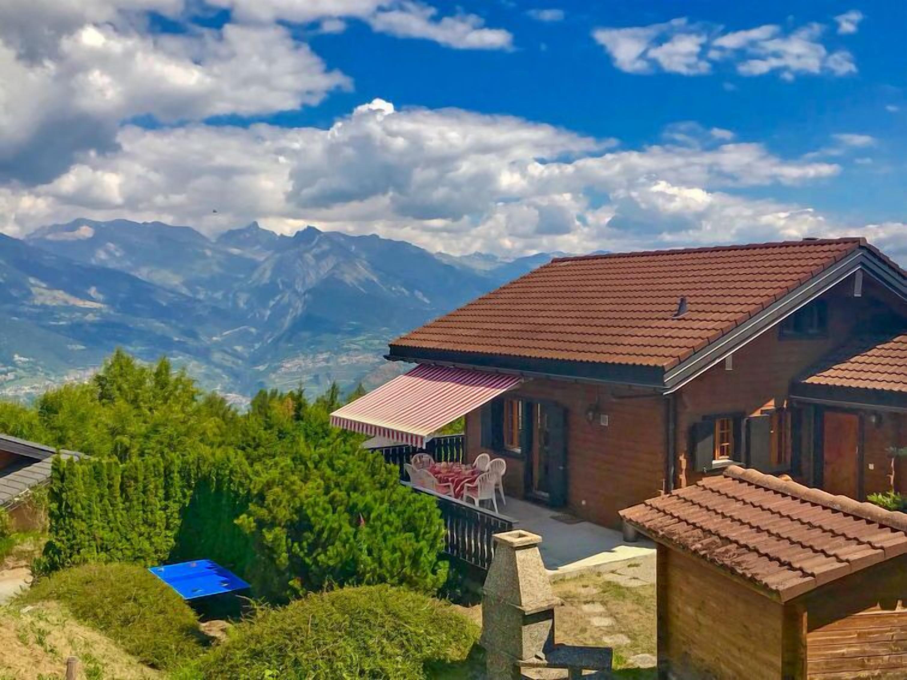 A chalet with a red-tiled roof, a patio with a striped awning, and a view of mountains in the background.