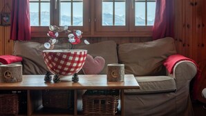 A cozy room with a wooden interior, a large window with curtains, a brown sofa, a table with a checkered bowl, pine cones, and two candles. - Vacation home Bivak by Interhome (Nendaz)