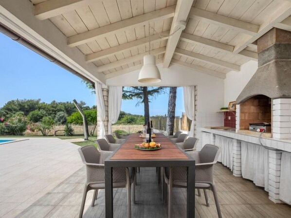 An outdoor dining area with a long wooden table and chairs, a stone fireplace, and a view of the surrounding landscape.