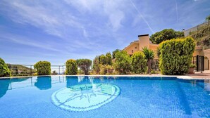 Una piscina azul cristalina con un diseño circular en el centro, rodeada de exuberante vegetación y un edificio de fachada color crema.