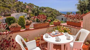 Una terraza con una mesa redonda y sillas blancas, rodeada de plantas en macetas y una vista panorámica del mar y las colinas.