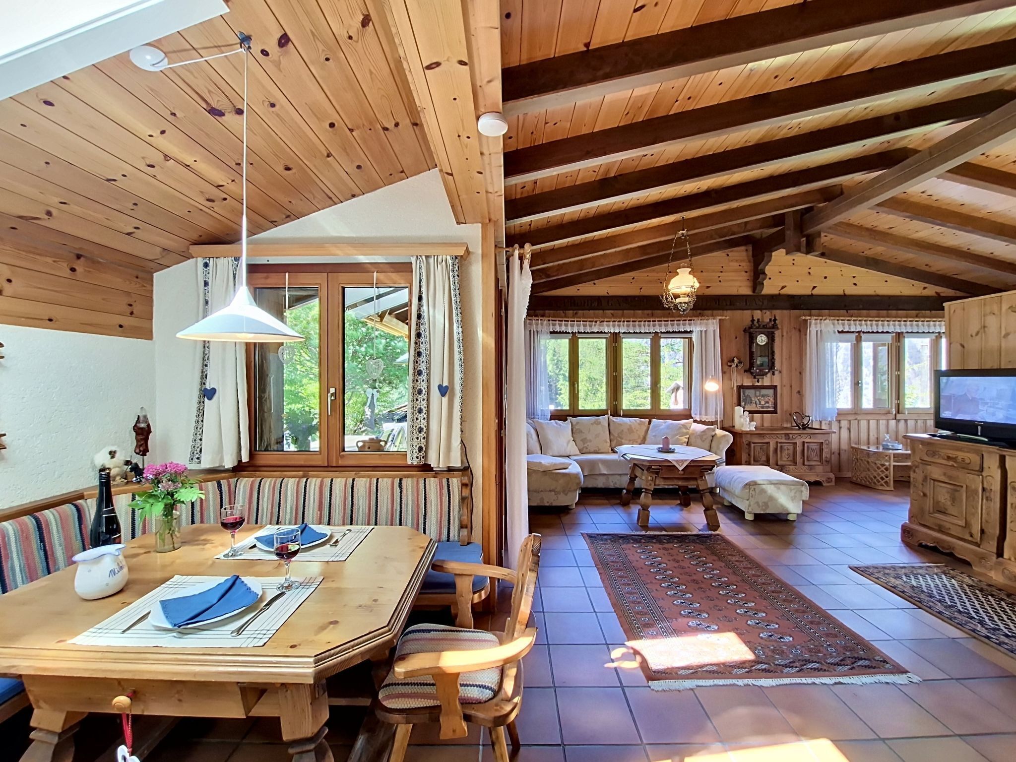 A wooden cabin interior with a dining area, sofa, TV, and a large window.