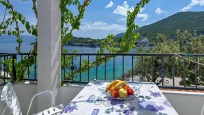 Un balcon avec une table dressée pour le petit-déjeuner, offrant une vue panoramique sur la côte, avec des montagnes et une étendue d’eau.