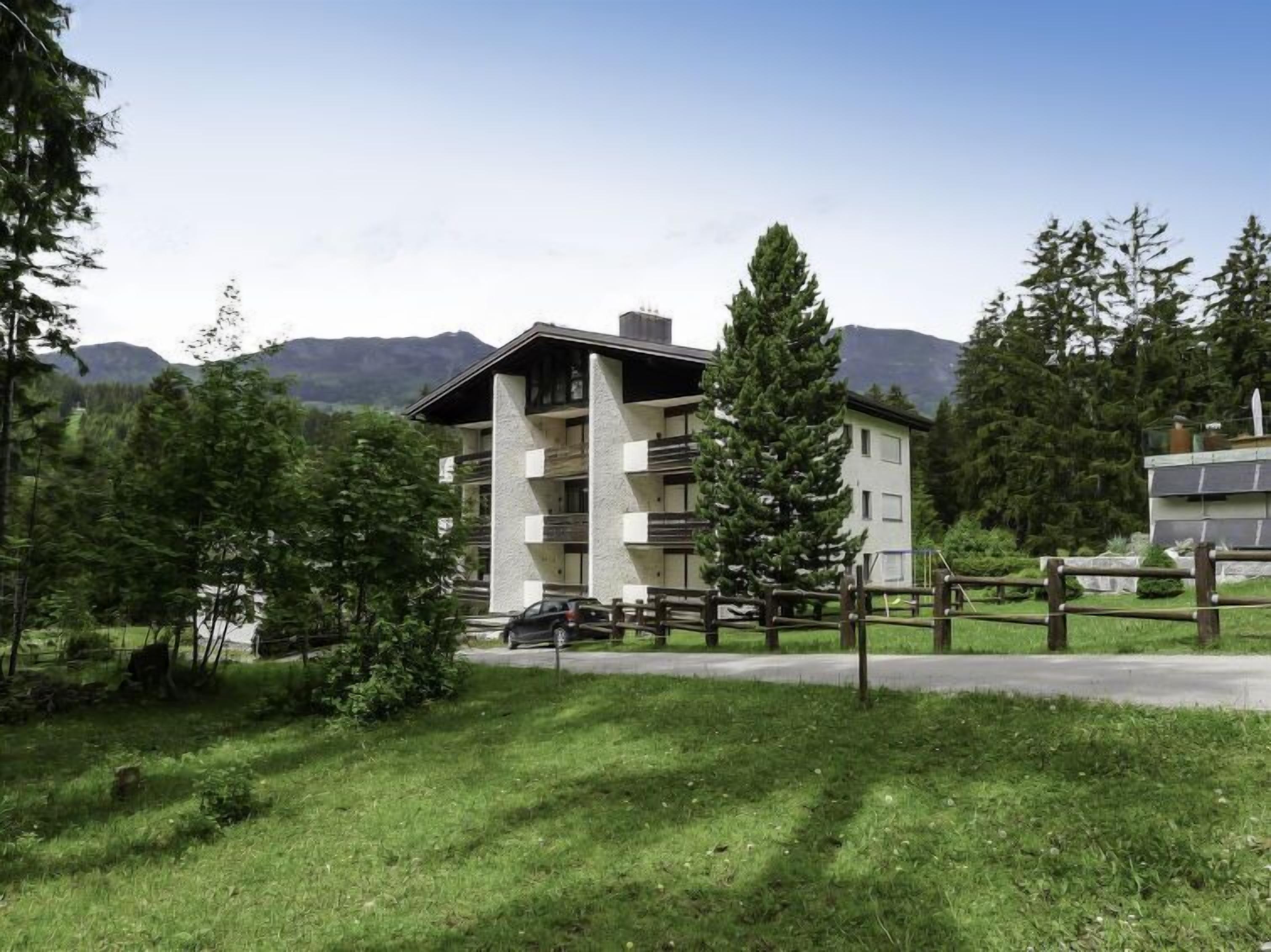 Un immeuble à plusieurs étages, doté de balcons, entouré de verdure et avec des montagnes en arrière-plan.