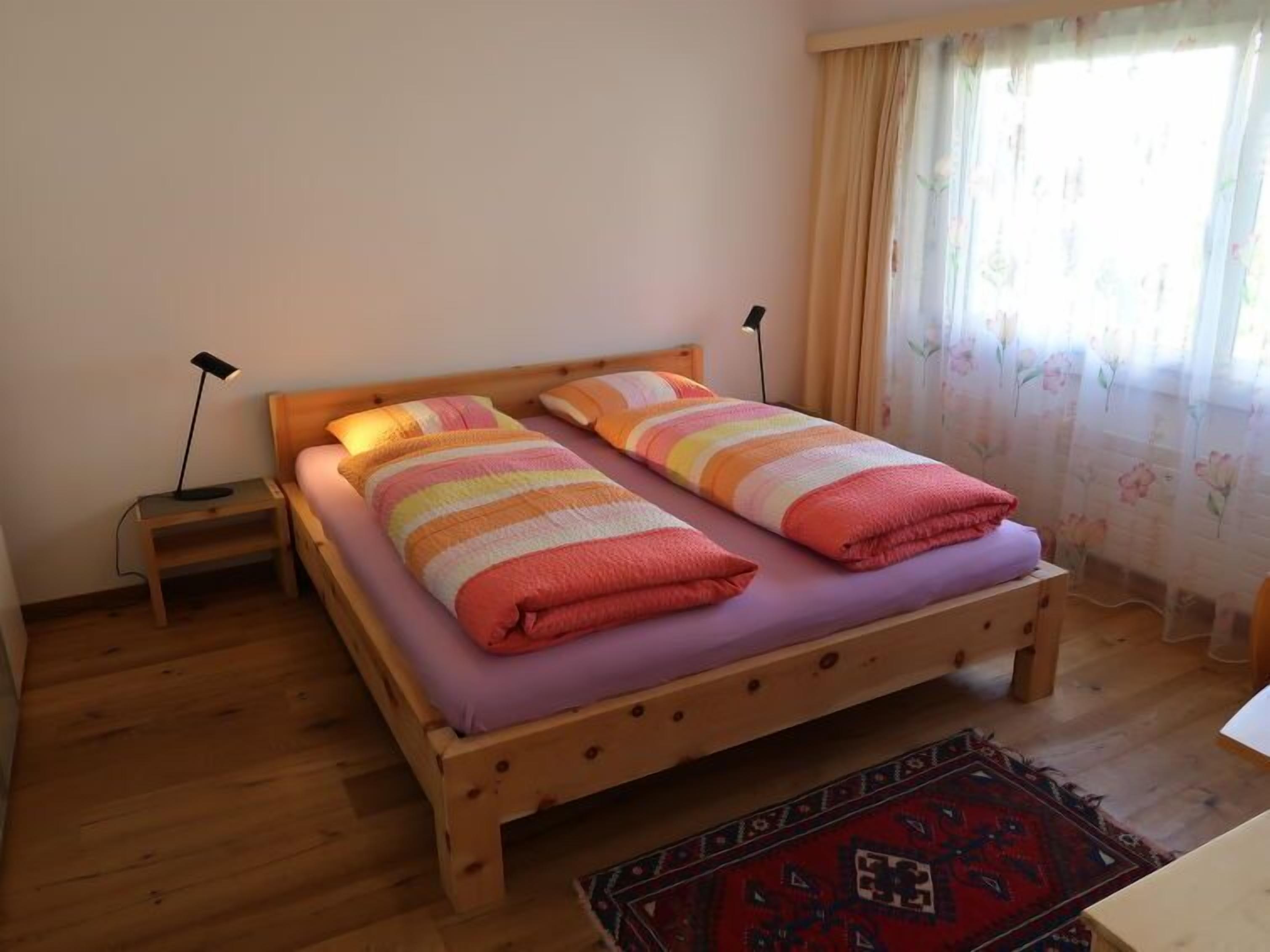 A single bed with a wooden frame, two pillows, a striped blanket, and a small bedside table with a lamp.