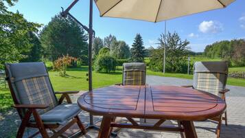 A patio with a wooden table and two chairs, a large umbrella, and a garden view.