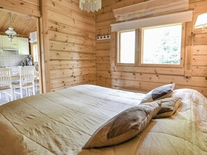 Quarto de cabana de madeira com uma cama, uma janela e área de cozinha visível através de uma porta aberta.