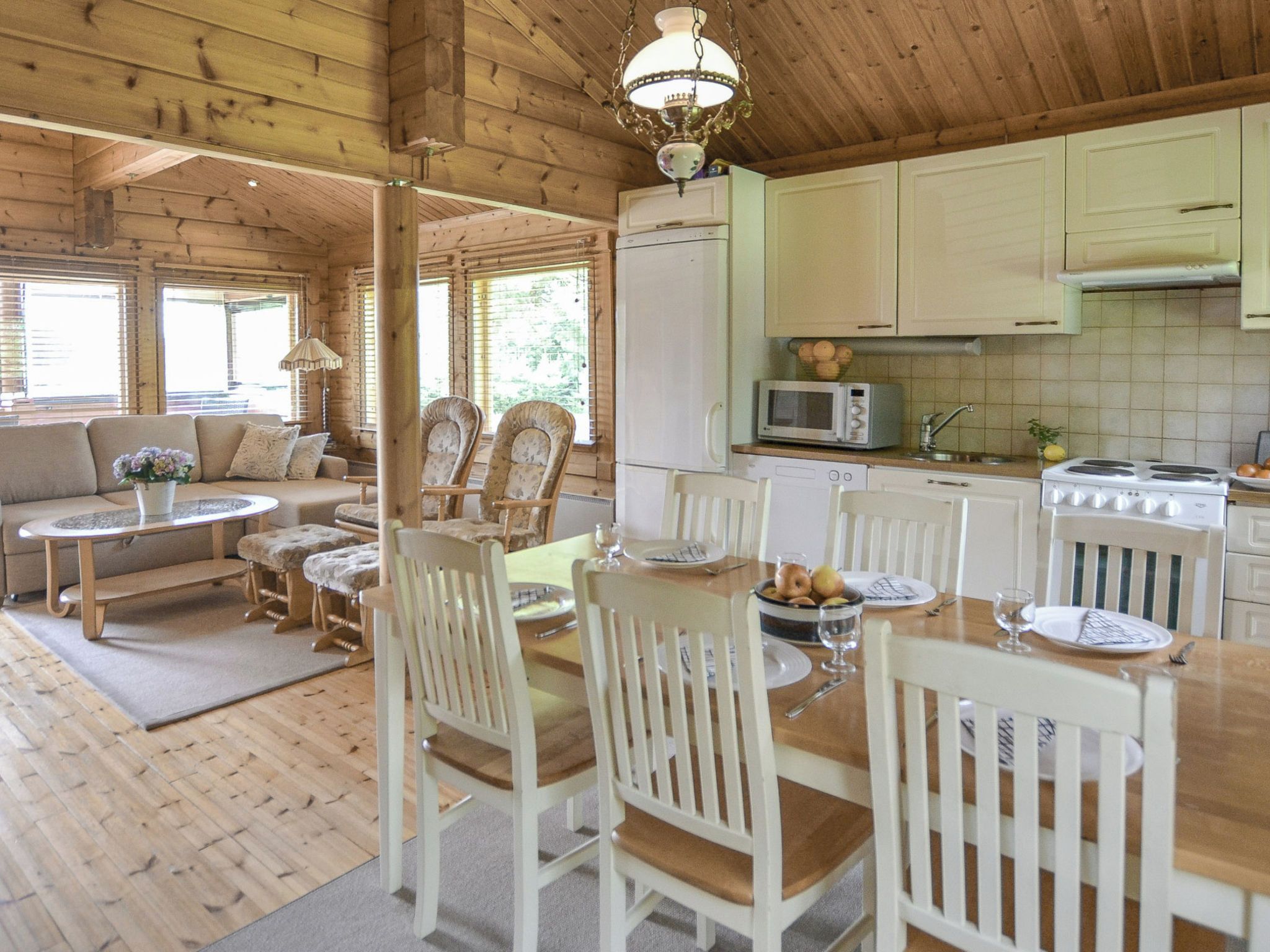 A wooden cabin interior with a dining area, kitchen, and a living room with a sofa and chairs.