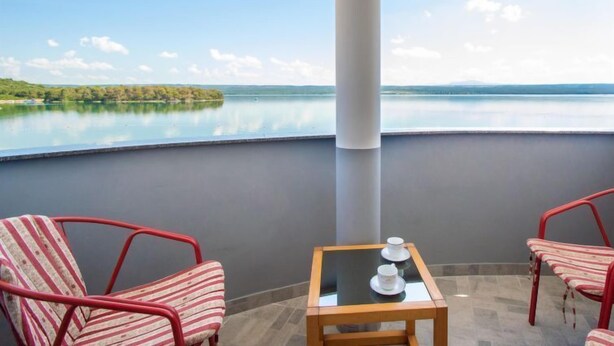 Ein Balkon mit Blick auf einen See und ferne Hügel, ausgestattet mit gestreiften Stühlen und einem kleinen Tisch mit Tassen.