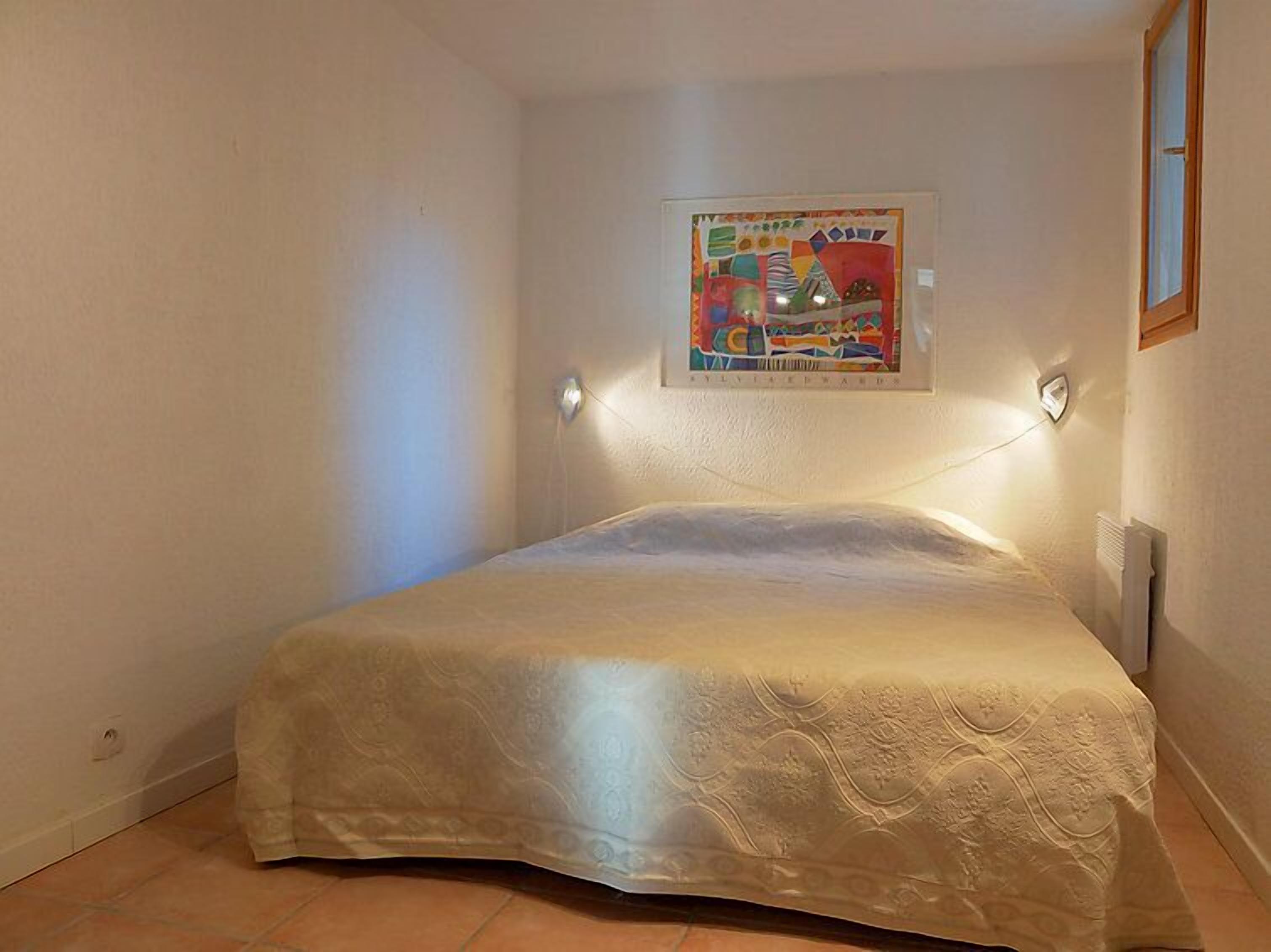 A small, neatly made bed in a room with white walls and a single framed artwork above it.