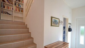 Un escalier recouvert de carreaux beiges, un banc en bois, une bibliothèque, un tableau encadré et un porte-manteau avec un sac.