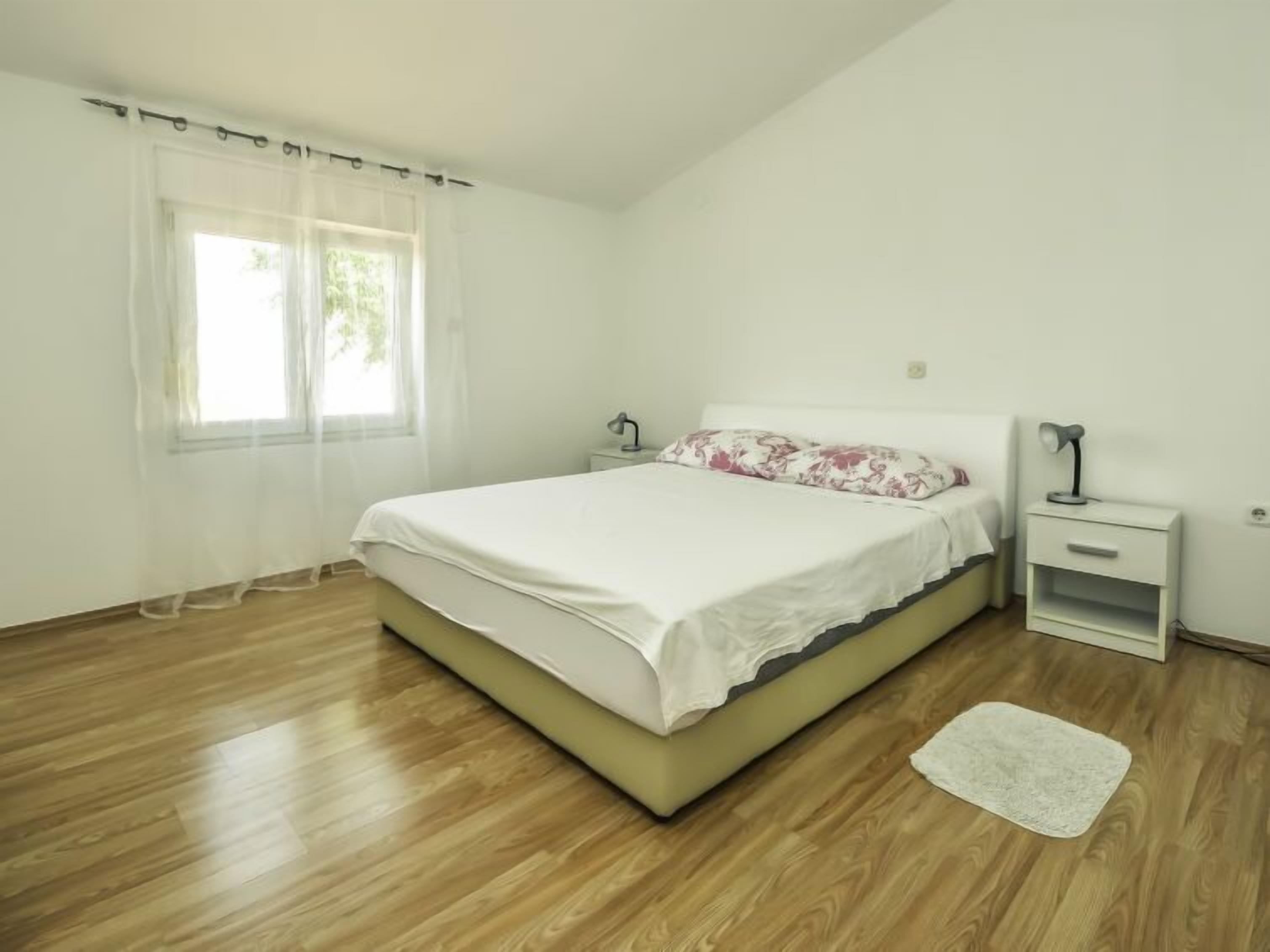 Une chambre à coucher avec un lit, une table de chevet, une lampe, une fenêtre avec des rideaux légers et un plancher en bois.