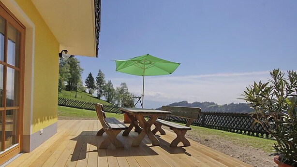Een houten terras met een tafel en stoelen, een groene parasol en uitzicht op een glooiend landschap.