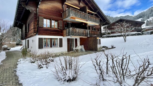 A wooden chalet with a balcony, a covered porch, and a snow-covered landscape.