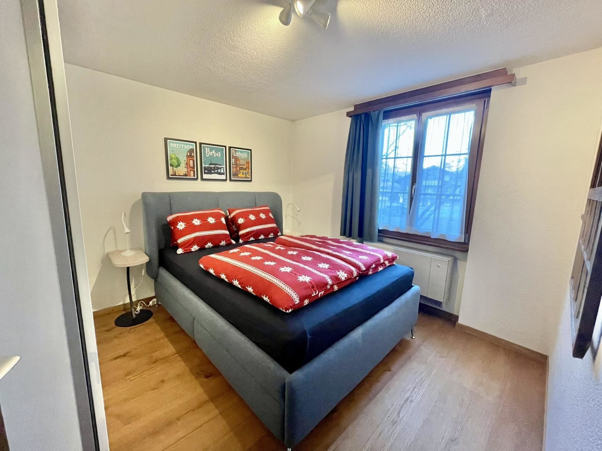 Une chambre à coucher avec un lit, une fenêtre avec des rideaux, un radiateur et des tableaux encadrés accrochés au mur.