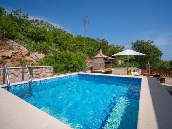 Uma piscina retangular com água azul cristalina, rodeada por um muro de pedra e uma escada de metal.