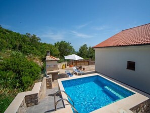 Piscina com um deck de pedra, área de estar com um guarda-sol branco e um edifício com telhado de telhas vermelhas.