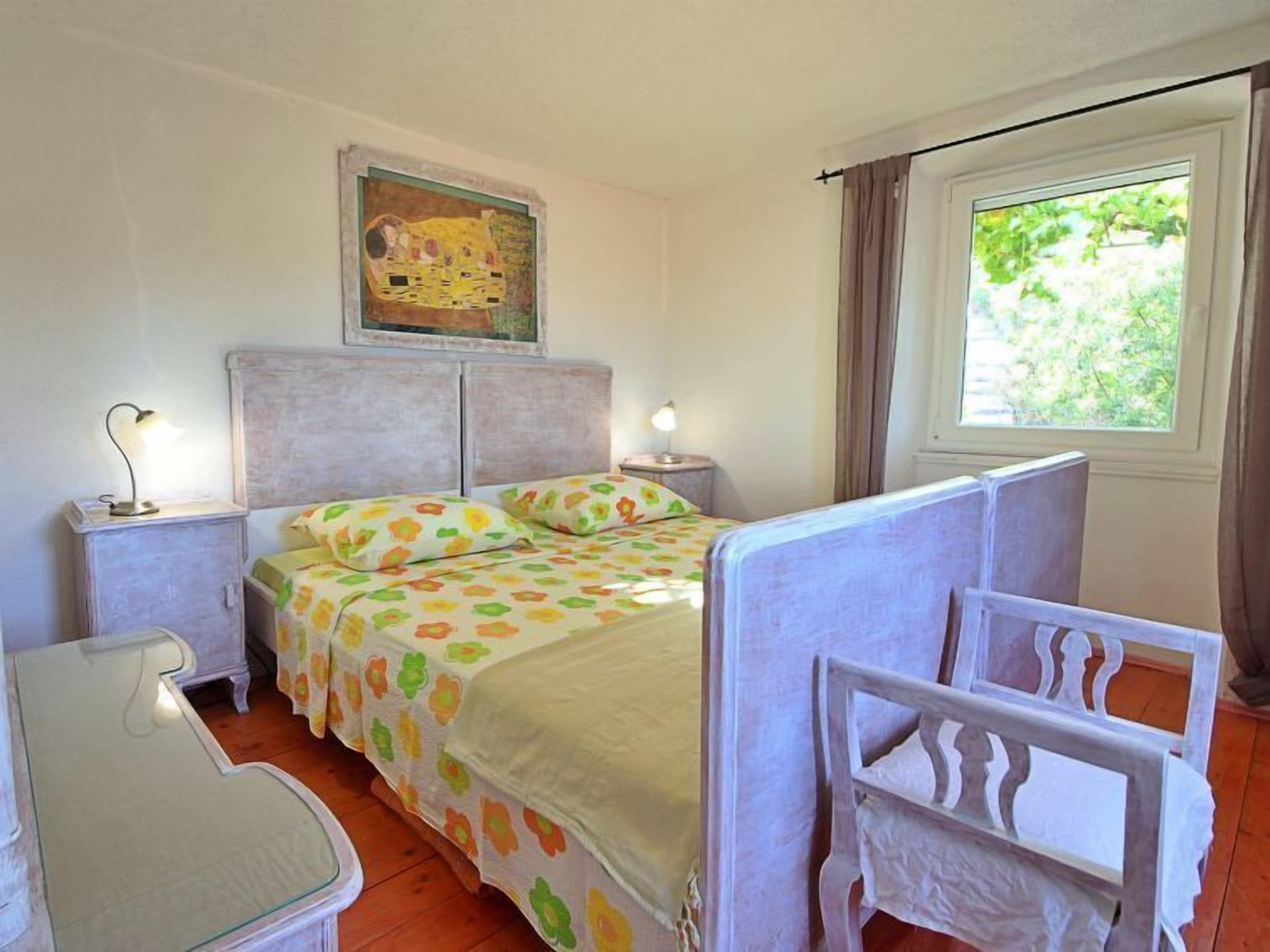 A bedroom with a bed, two bedside tables, a chair, a painting on the wall, and a window with curtains.