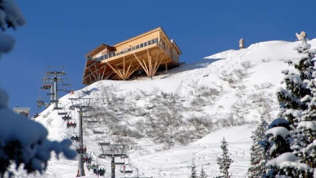 Un chalet en bois situé sur une pente enneigée, avec des remontées mécaniques en arrière-plan.