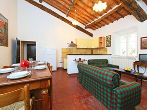 Una zona d'estar acollidora amb taula de dinar de fusta per a quatre, un sofà amb quadres verds i vermells, una cuina amb armaris de fusta, i una finestra amb vistes a vegetació.