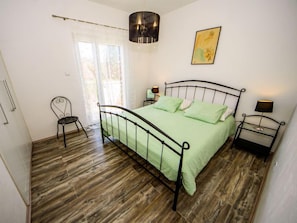 A bedroom with a metal bed frame, a green bedspread, two bedside tables with lamps, a chair, and a window with sheer curtains.