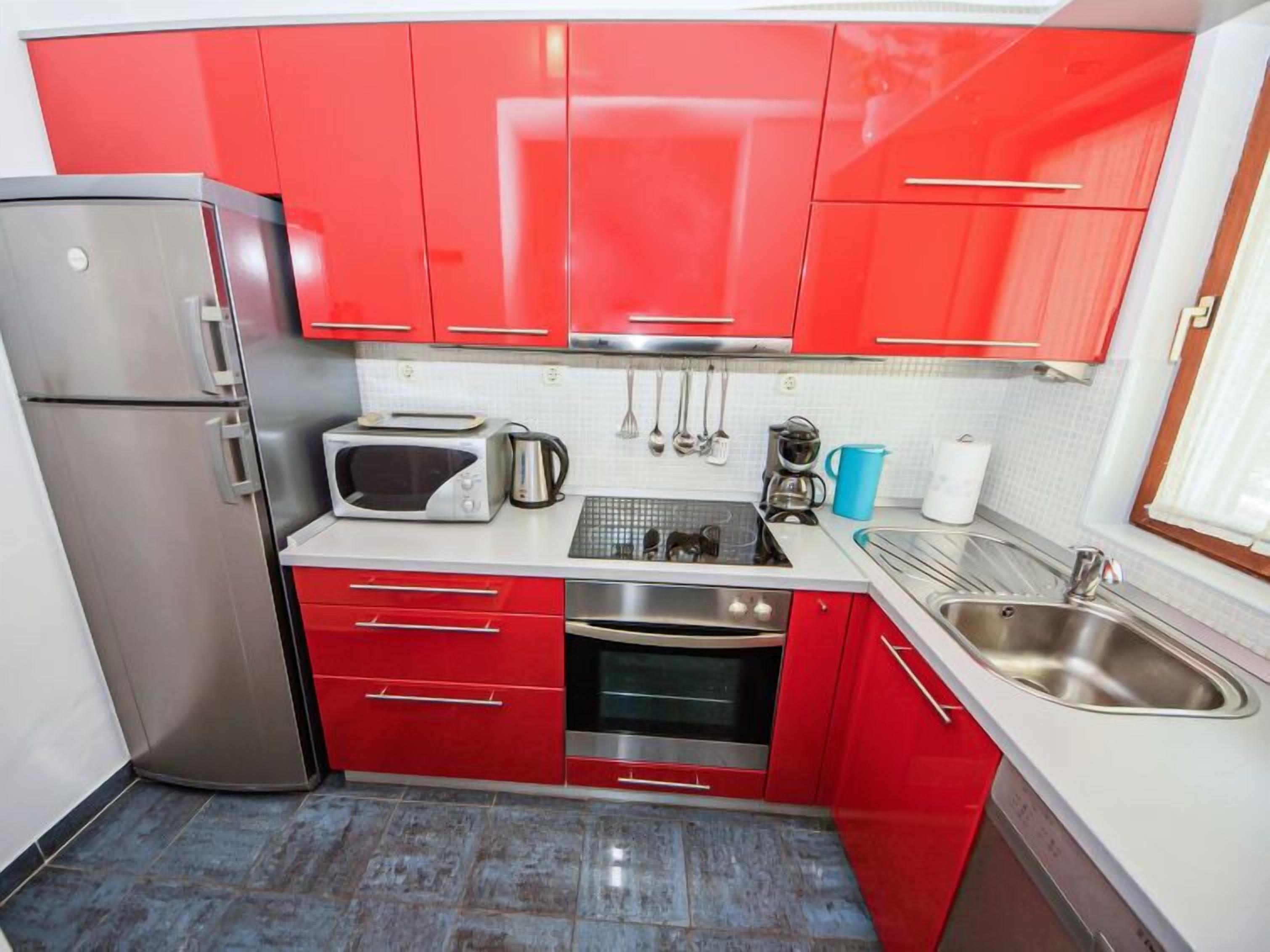 Una cocina compacta con armarios rojos, un refrigerador de acero inoxidable, un microondas, una tetera, una cafetera y un lavamanos.