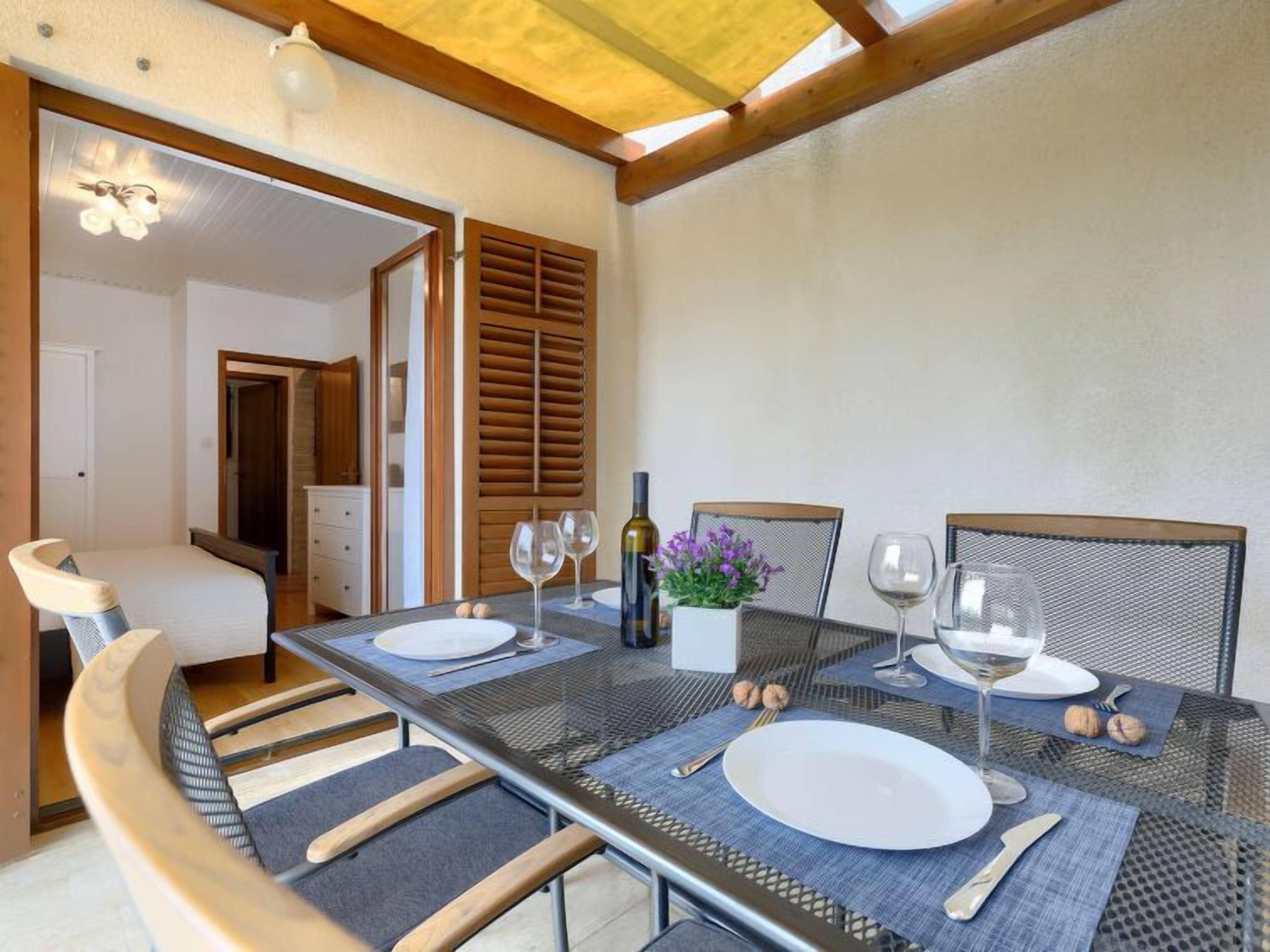 Un coin repas avec une table dressée pour deux personnes, une chaise et une petite table sur laquelle se trouvent une bouteille et des fleurs.