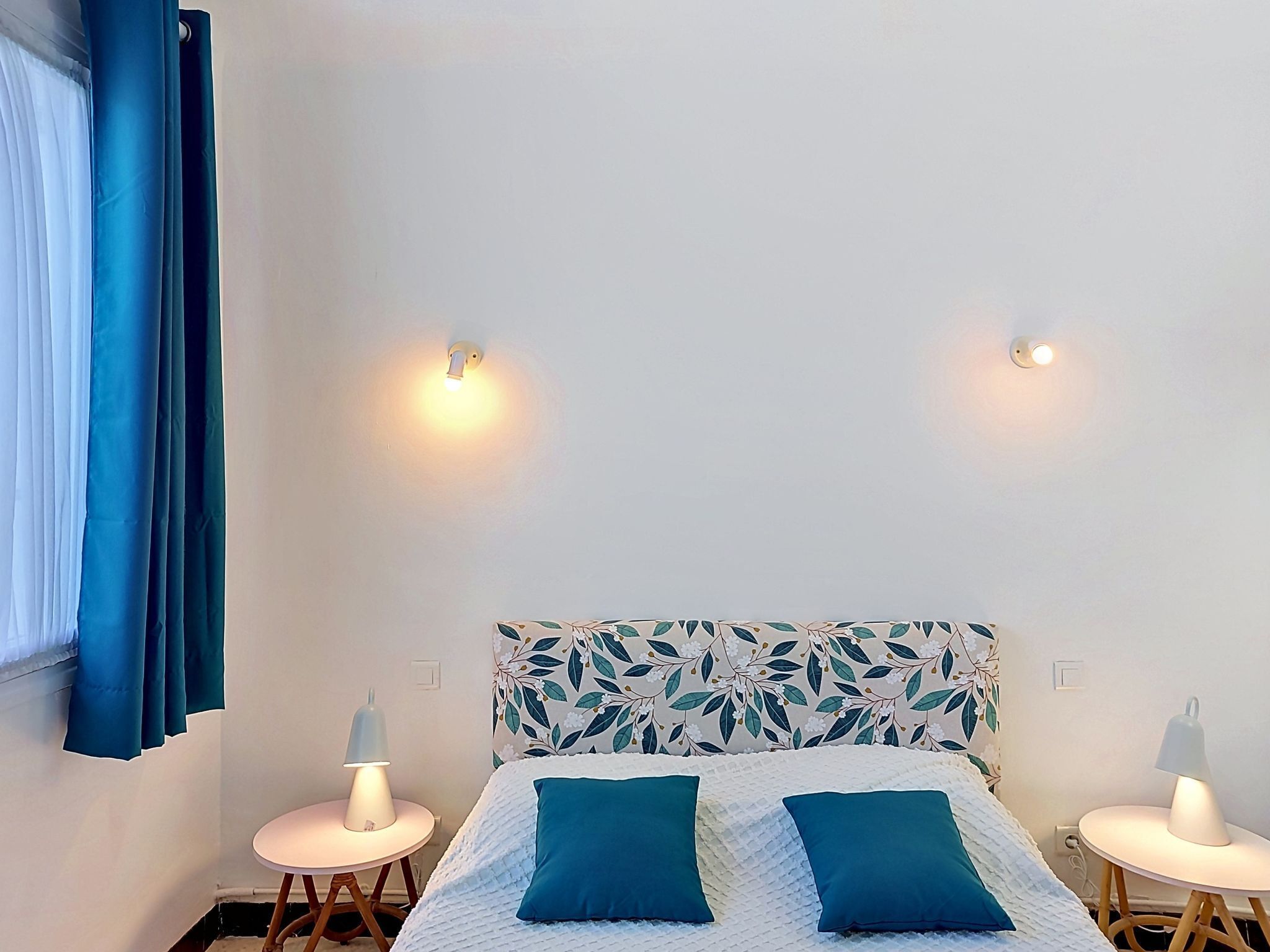 A bedroom with a bed, two bedside tables, a headboard with a floral pattern, and two wall-mounted lights.