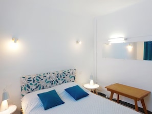 A bedroom with a bed, two pillows, a bedside table, a lamp, a mirror, and a wall-mounted light fixture.