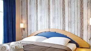 Ein Hotelzimmer mit einem hölzernen Kopfteil, einem Bett mit weißer und blauer Bettwäsche, einem Nachttisch mit einer Lampe und einem Fenster mit blauen Vorhängen.