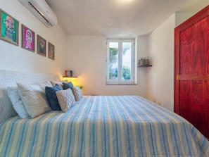 A bedroom with a bed, pillows, a window, a shelf with books, and a red door.