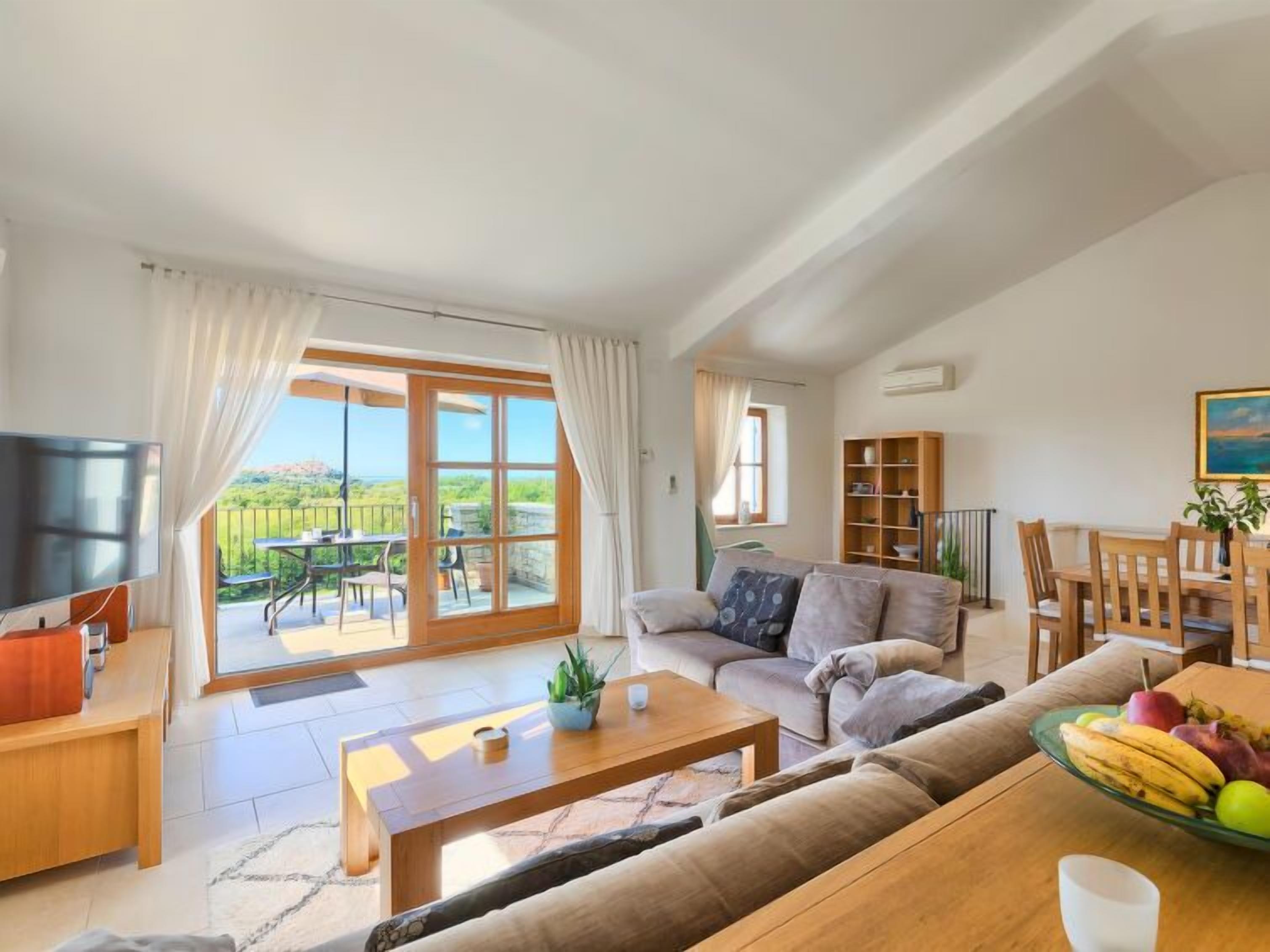 Uma sala de estar espaçosa com uma grande porta de vidro deslizante que dá para uma varanda, uma televisão, uma mesa de centro de madeira, um sofá e um espaço de jantar com uma fruteira.