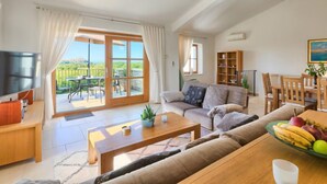 Een ruime woonkamer met een grote schuifpui naar een balkon, een televisie, een houten salontafel, een bank en een eethoek met een schaal fruit.