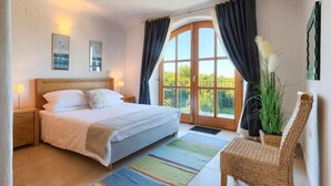 Een slaapkamer met een bed, nachtkastjes, een stoel, een raam met gordijnen en uitzicht op groen.