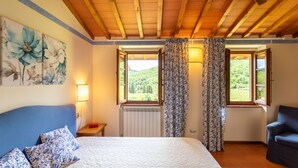 Een slaapkamer met een groot bed, een stoel, een radiator en twee ramen met gordijnen.